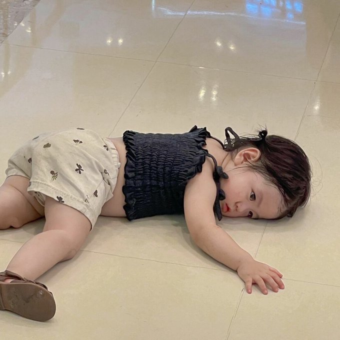 A baby wearing a black top and cream-colored shorts with small patterns lies on a tiled floor, looking toward the camera with one arm extended.