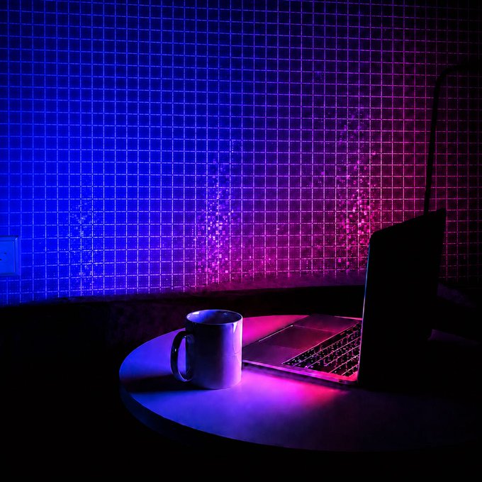 A laptop and coffee mug sit on a round table bathed in moody neon blue and pink light against a grid-tiled wall, evoking a late-night solo work session — fitting the "one-person company with a night team" vibe, since that "team" is you, fueled by caffeine after hours.
