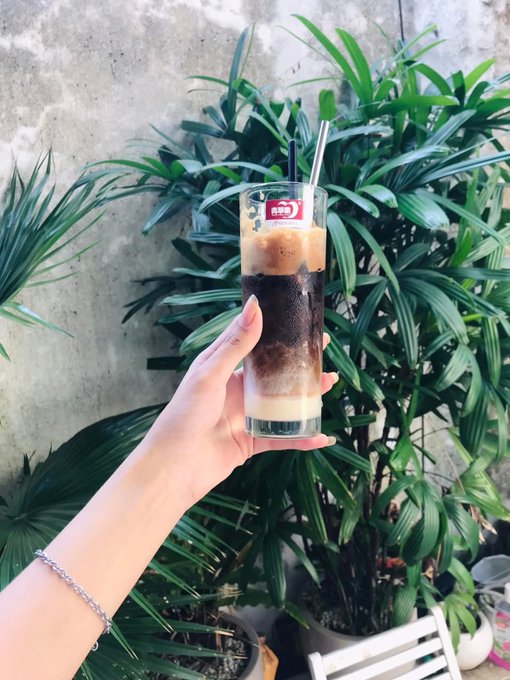 A hand holding up a tall layered iced coffee drink with a red logo sticker, photographed against a background of green palm-like plants.