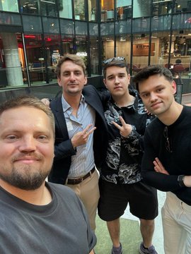 The image features four young men posing for a photo together, with one of them wearing sunglasses. They are standing outside a building, possibly a restaurant or bar, as indicated by the presence of a car visible in the background. The group appears to be enjoying their time and is likely friends who have gathered for a casual outing or event.