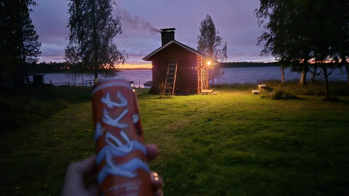A hand holds a red 'JOKA' energy drink can in the foreground, with a small wooden cabin by a lake at sunset in the background, featuring a ladder on the cabin and a smokestack.