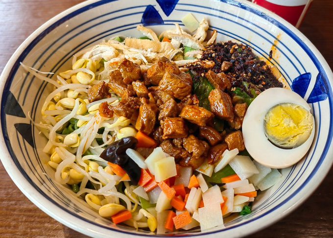 A vibrant bowl of Taiwanese-style noodle soup featuring braised pork, bean sprouts, diced vegetables, chili oil, and a halved boiled egg, served in a blue and white patterned bowl, presented as a delicious afternoon meal.