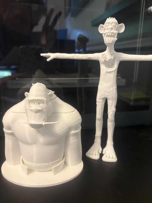 Two 3D-printed white figurines on a dark surface: a rotund, seated character with prominent teeth on the left, and a tall, thin monkey-like character in a T-pose on the right.
