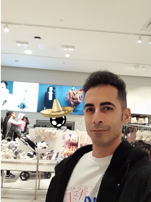 A selfie of a man in a white t-shirt and dark jacket inside a retail store, with a sombrero placed on what appears to be a ball in the background.