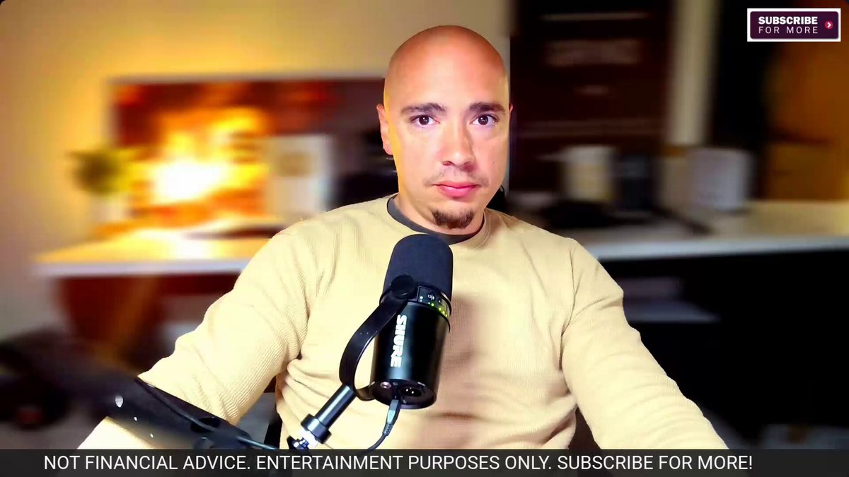 The image features a bald man wearing a beige sweater sitting behind a microphone with his hands resting on his knees. The background of the image shows a desk, suggesting that he might be at work or in an office setting. There are also some books visible near him, which could indicate that he is involved in a profession related to finance or business.