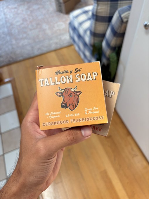 A hand holds an orange box of 'Health y Sol' 'TALLOW SOAP' labeled 'Cedarwood Frankincense', '5.5 oz bar', 'All Natural Organic', and 'Grass-Fed & Finished', with a cow illustration.