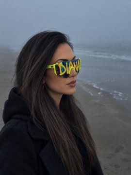A woman with long dark hair stands on a foggy beach wearing bright neon yellow sunglasses with the word 'TDIANA' written across them.