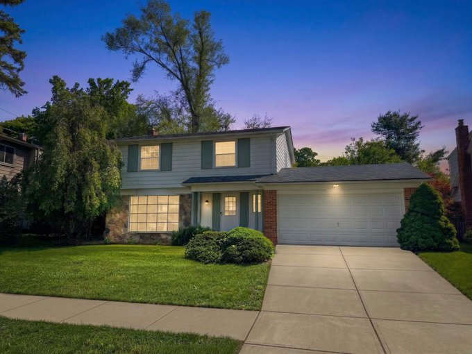 The image shows a house with a garage door at dusk. The house has a large lawn surrounding it, and there are trees visible nearby. In front of the house, there's a sidewalk leading up to the entrance. The scene appears to be captured during sunset or twilight hours when the sky is filled with warm hues from the setting sun.