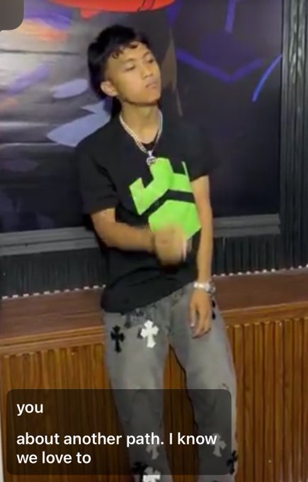 A young man wearing a black t-shirt with a green logo and jeans with cross patches stands indoors. A text overlay reads 'you about another path. I know we love to'.