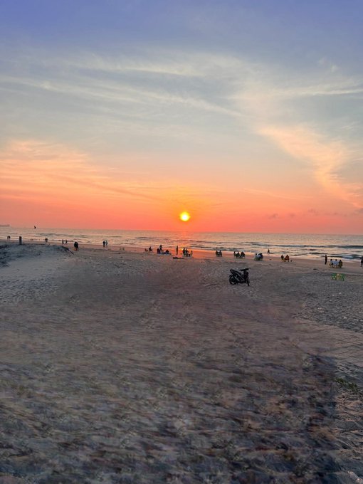 A serene beach scene at sunset, with the sun low on the horizon casting orange and pink hues across the sky, while silhouettes of people can be seen scattered along the shoreline.