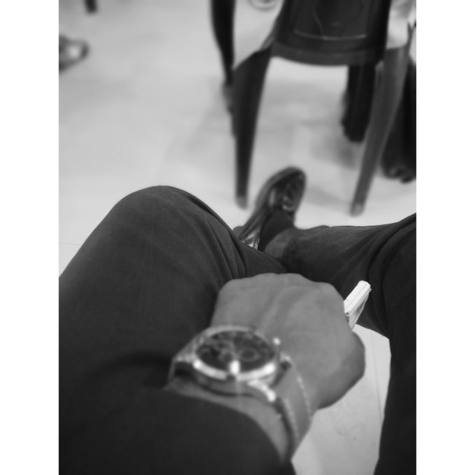 A black and white first-person perspective photo showing a person's lap with dark jeans and a wristwatch, while holding a pen, with another person visible in the background.
