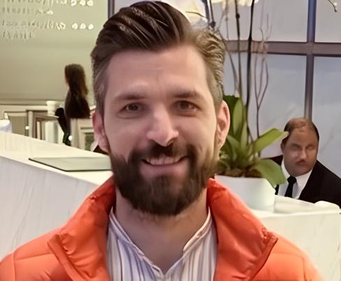 A bearded man in an orange jacket smiles at the camera in the foreground, while a man with a mustache in a suit stares suspiciously from the background, resembling the 'Distracted Boyfriend' meme format.