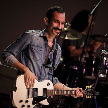 The image features a man playing an electric guitar, smiling as he performs. He is wearing a blue denim shirt and has his arms around the neck of the guitar. In the background, there are two other people present - one person is holding a drum set, while another person is standing behind them. The post that reads "My artistic range" suggests that this image captures an interesting moment where multiple musical instruments come together to create a unique and creative performance.

The man playing the electric guitar appears to be enjoying himself as he plays in front of his friends. His smile indicates that he is having a good time, showcasing his talent and passion for music. The presence of other musicians in the background adds depth to the scene, emphasizing the collaborative nature of their musical endeavors.