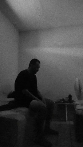 The image shows a man sitting alone in a room with his head resting on a pillow or cushion. The room appears to be dimly lit, creating an atmosphere of solitude and contemplation. The man's posture suggests that he may be deep in thought or experiencing some form of emotional distress. In the background, there is a TV screen mounted on the wall, which could potentially be used for entertainment purposes during his time alone.