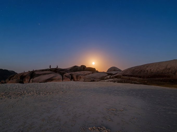 A serene twilight landscape showing the moon rising over rocky terrain with a few silhouetted figures standing on the boulders.