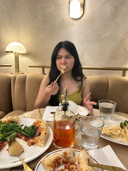 A woman with long dark hair eats pizza with a golden fork at a restaurant table filled with pizza slices, pasta, salad, an olive oil dispenser, and water glasses.