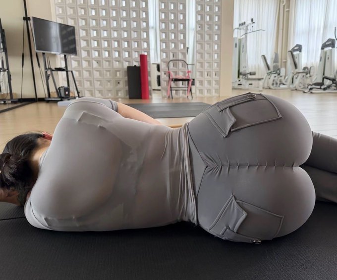 A person lying face-down on a yoga mat in a gym, wearing a sheer light-colored long-sleeve top and tight beige cargo pants, with exercise equipment visible in the background.