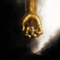A close-up image of a golden pendant or medallion featuring a hand design, illuminated dramatically against a dark background.