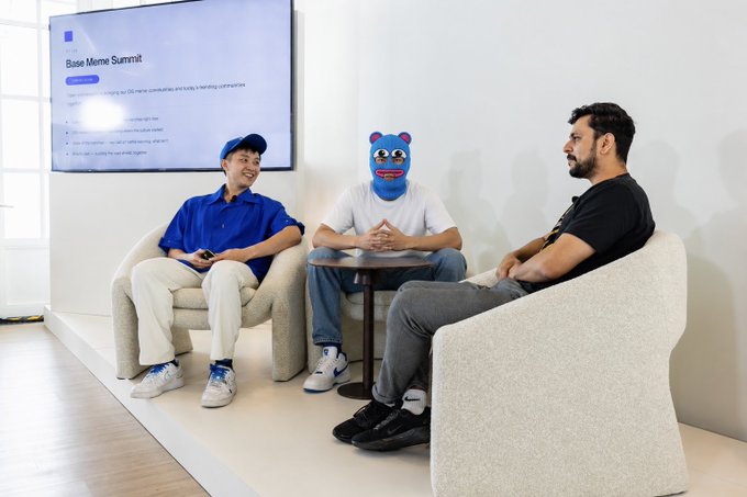Three people sit in a bright studio-style meeting area; the central person wears a blue cartoon mask, and a wall display reads 'Base Meme Summit.'
