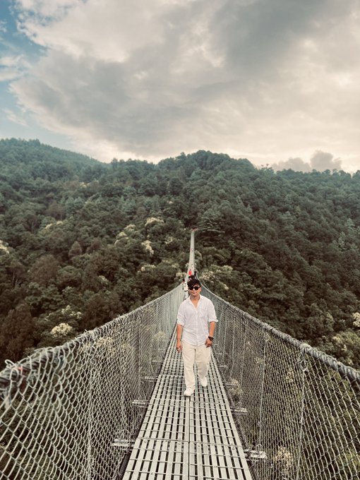 A man in a white shirt and beige pants walks across a long suspension bridge over a lush, green forested valley under a cloudy sky.