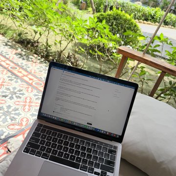 A laptop displaying code or an AI interface rests on a cushioned outdoor chair, with a coffee cup nearby and a garden in the background. The scene captures a serene Saturday morning workflow, blending nature, caffeine, and AI tools (Claude/Codex and OKX AI) for organizing thoughts and bringing ideas to life.