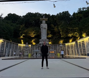 A person stands in the center of a cemetery courtyard, posing in front of a large statue with a hill of illuminated graves and niches behind.