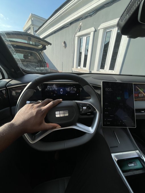 A first-person view from the driver's seat of a car shows a hand on the steering wheel, with digital dashboard displays showing speed and range, parked outside a building.