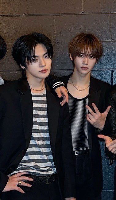 Two stylish young men pose together against a gray cinderblock wall, both wearing black blazers over striped tops; the one on the left has dark hair and a striped shirt with a silver ring on his finger, while the one on the right has light brown hair, wears a striped tee, and flashes a peace sign.