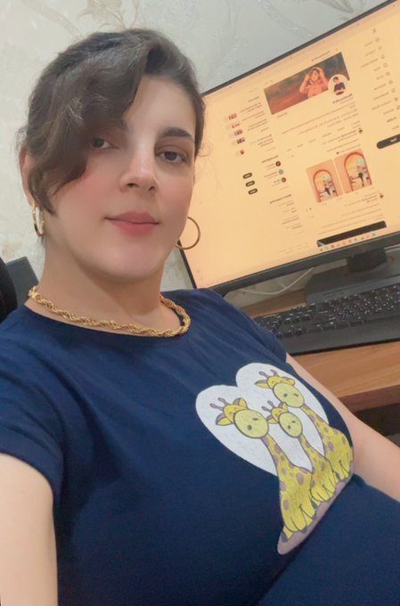 A selfie of a woman with short dark hair wearing a dark blue t-shirt featuring a graphic of three giraffes, sitting in front of a computer monitor.