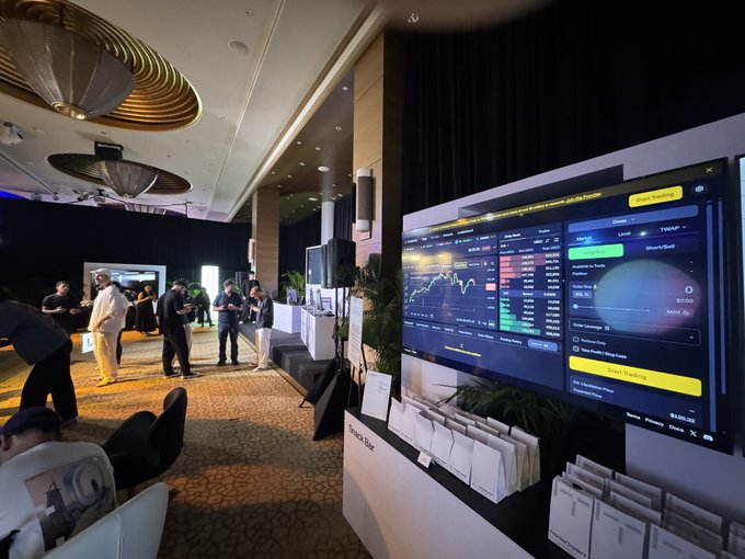 A photo from a financial or cryptocurrency conference showing attendees near a 'Simple Bot' booth with a large screen displaying trading interfaces and market data.
