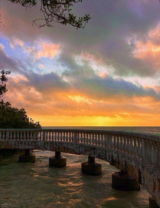 A scenic photograph of a stone bridge or pier at sunset, with dramatic clouds and golden light reflecting on the water below.