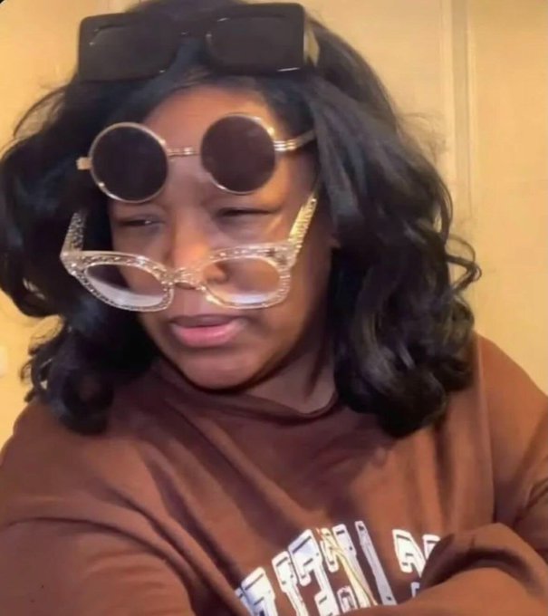 A woman wearing a brown sweatshirt, with sunglasses perched on her head and two additional pairs of round glasses hanging down over her face, posing for a selfie.
