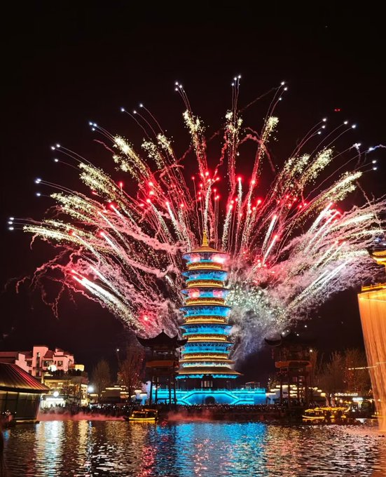 A spectacular nighttime fireworks display erupts over an illuminated multi-tiered pagoda tower reflected in the water below.