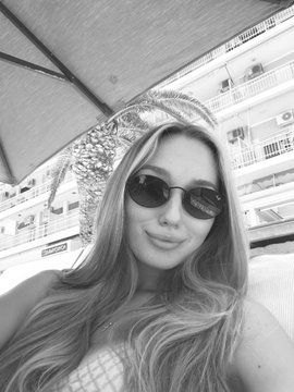 A black-and-white selfie shows a woman wearing sunglasses and a sun hat, with city buildings and an umbrella in the background.