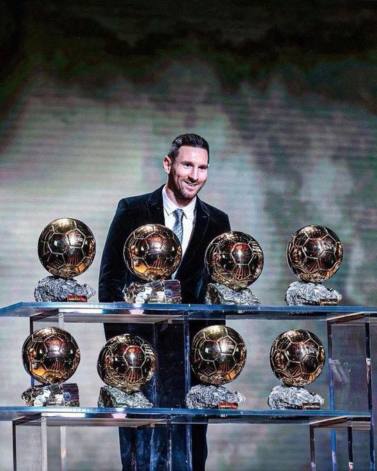 The image features a man standing next to a table with several trophies displayed on it. The man appears to be smiling as he looks at the trophies, possibly celebrating his achievements or accomplishments. The trophies are arranged neatly on the table, showcasing various sizes and shapes. The context of the image suggests that these trophies may represent awards for different competitions or achievements in sports, academics, or other fields.
