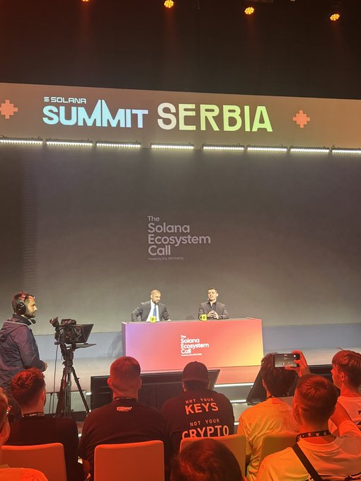 Two men seated on stage at "SOLANA SUMMIT SERBIA" during "The Solana Ecosystem Call" presentation, with attendees in the foreground, one wearing a shirt reading "NOT YOUR KEYS NOT YOUR CRYPTO."