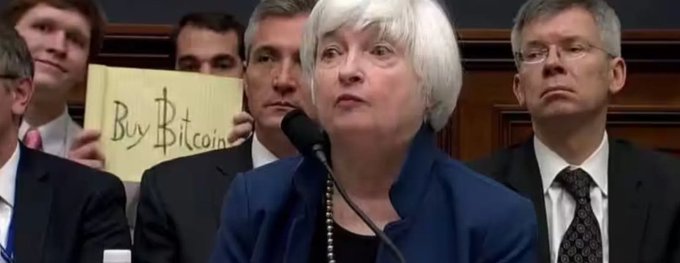 Janet Yellen speaking at a podium while a person behind her holds up a yellow sign reading 'Buy Bitcoin'.