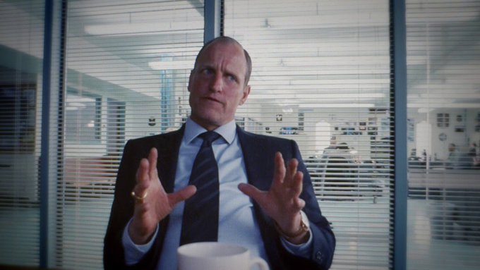 A scene from a film or TV show showing a man in a suit gesturing with his hands while sitting in an office with venetian blinds.