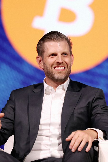 A smiling man with short brown hair and a beard, wearing a dark suit jacket and white shirt, sits in front of a backdrop featuring a large Bitcoin (₿) logo. The image relates to the accompanying post about Eric Trump, who is pictured here, discussing how AI, stablecoins, and tokenization could accelerate cryptocurrency adoption in everyday finance.