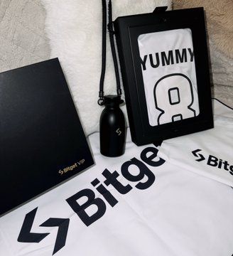 A Bitget branded merchandise bundle laid out on a bed, including a black box, a water bottle with lanyard, a framed 'YUMMY' print, and a white t-shirt with the Bitget logo.
