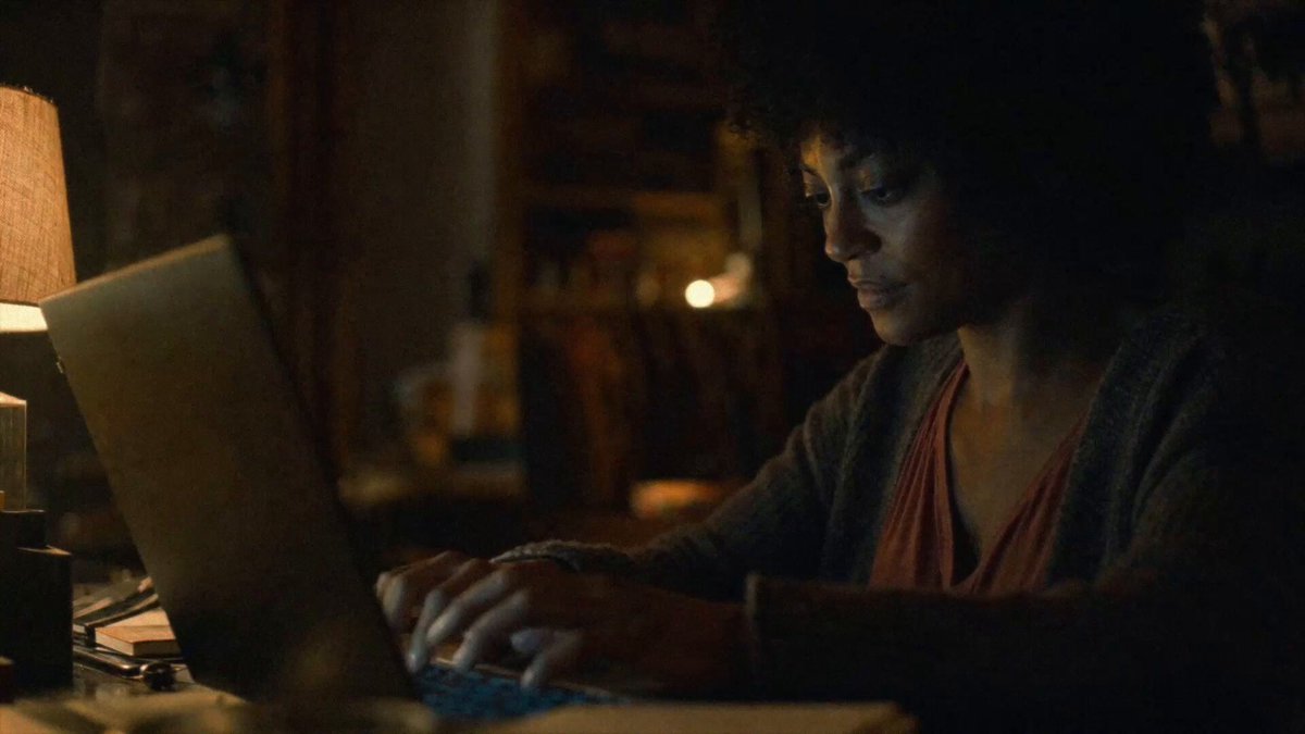 The image shows a woman sitting at a desk, typing on her laptop computer. She has curly hair and is wearing a gray sweater. In front of her are several books, suggesting that she might be reading or studying while working on her laptop. The background appears to be a dark room with a lamp providing light for the scene. The image also includes text that reads "Solana is about to make a memory." This could be interpreted as a reference to the movie "The Dark Knight" where the character Solana has a memorable moment in the film.