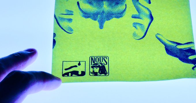 A hand holding a yellow-green print with blue ink illustrations and a 'NOUS' logo at the bottom center.