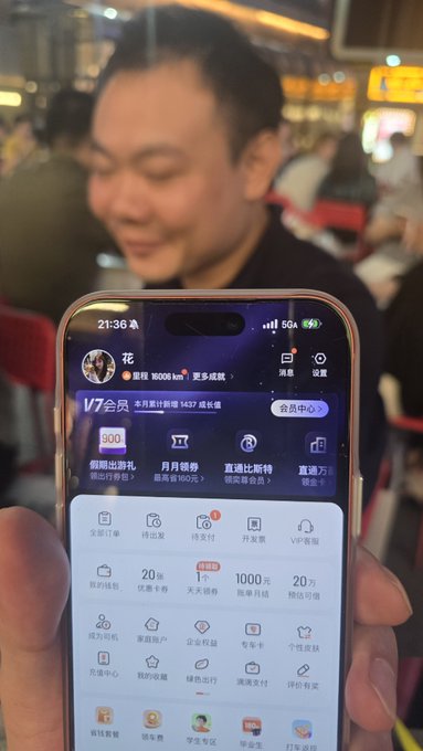 A man smiling in the background while holding up a smartphone in the foreground displaying a Chinese-language app interface with a 'V7会员' promotion and various service icons.