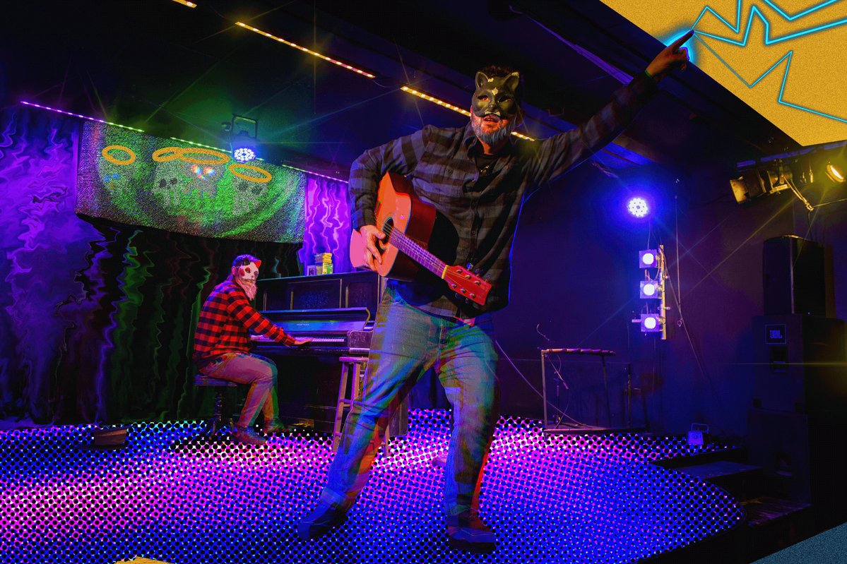 The image shows a man playing guitar while another person sits at a piano behind him. The scene takes place in front of a stage, which has purple lights illuminating the area. There are also speakers placed around the room, suggesting that this could be an event or performance taking place on stage.