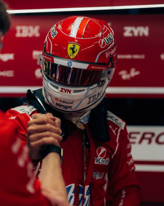 A Ferrari driver, identifiable as Charles Leclerc by the "Richard Mille" branding and prancing horse logo, stands in the pit garage wearing his iconic red racing suit and matching helmet adorned with sponsors including Shell, HP, ZYN, IBM, UniCredit, and BingX, while clasping hands with a teammate in a fist bump that embodies the bullish, performance-driven spirit heading into the Azerbaijan Grand Prix in Baku.