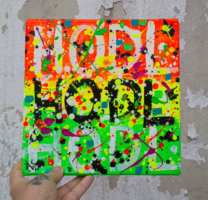 A hand holds a vibrant, abstract painting on a small canvas featuring the words 'YOU LOOK', 'GOOD', and 'FOOD' in large block letters, covered in colorful paint splatters.