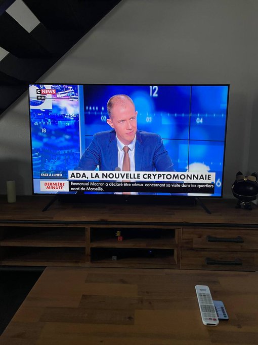 A television displays a French news broadcast from BFM TV with the headline 'ADA, LA NOUVELLE CRYPTOMONNAIE' about Emmanuel Macron's visit to Marseille, sitting on a wooden stand with a remote control nearby.