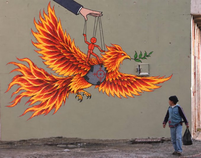Street art on a building wall depicting a fiery orange phoenix carrying a red figure controlled like a marionette, with a man in a blue sweater walking past on the sidewalk below.