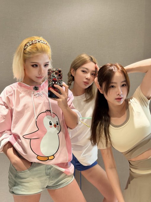 Three young women pose for a mirror selfie in casual outfits, with the woman on the left wearing a pink hoodie featuring a large penguin graphic.