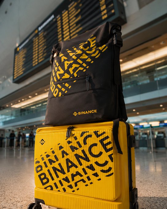 A black backpack featuring "BINANCE" branding rests atop a matching yellow suitcase, both displaying repeated "BINANCE" text, stacked in what appears to be an airport terminal with a flight information board in the background—fittingly capturing the "Off we go" sentiment of travel departure.